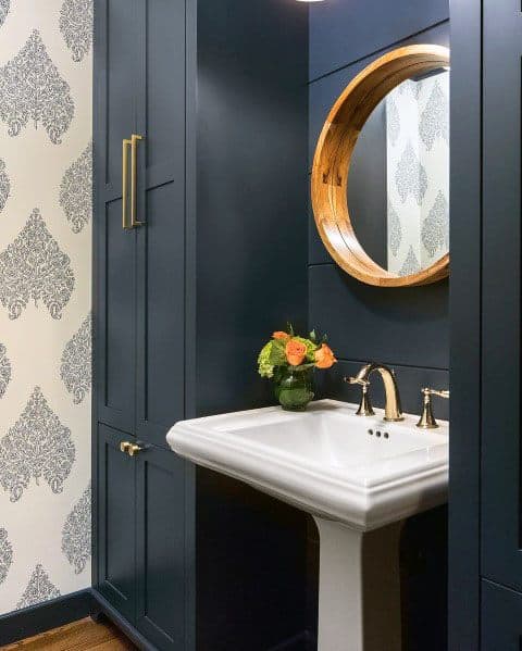 Blue bathroom with pedestal sink, round wooden mirror, and patterned wallpaper.