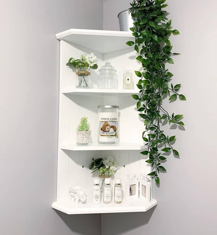 White corner shelf with greenery, candles, decorative jars, and small plants.