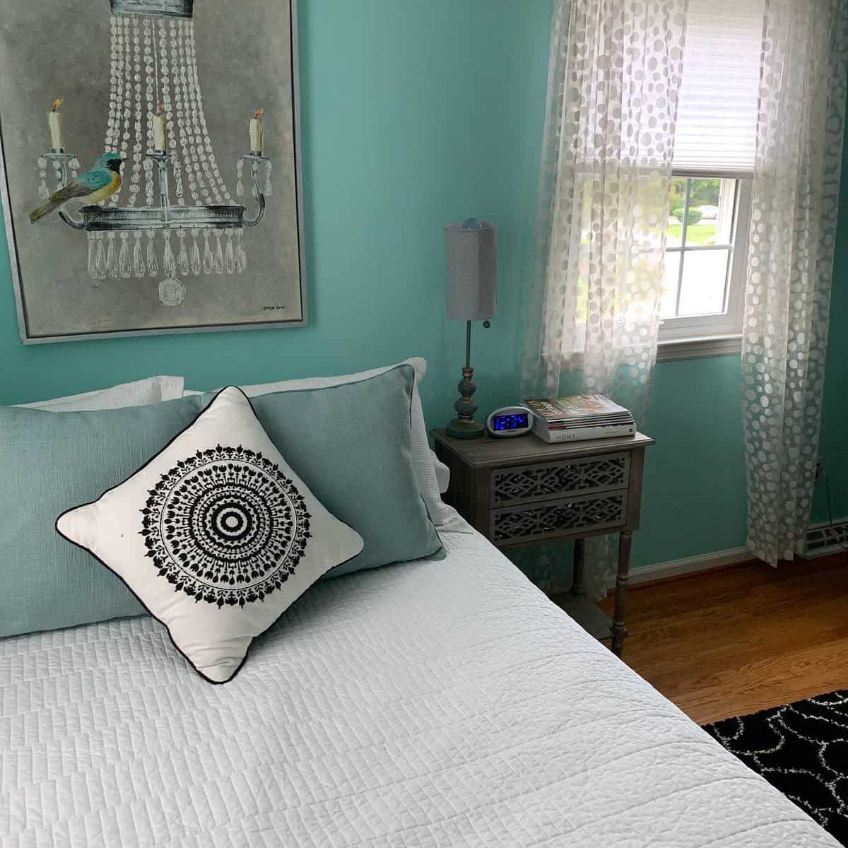 Bedroom with teal walls, bed with patterned cushions, side table with books, and decorative art on the wall