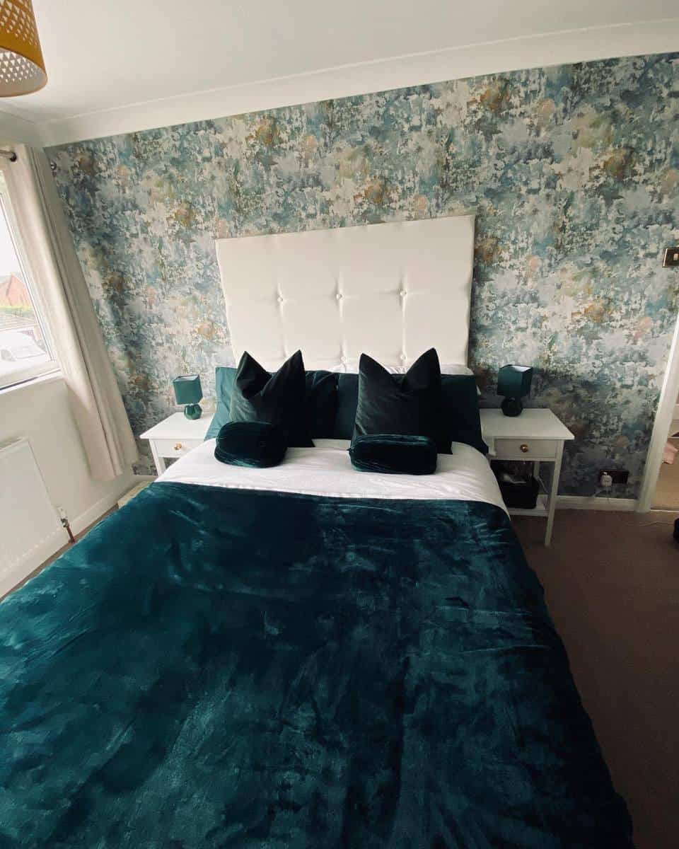 Bedroom with a large white headboard, teal bedding, two side tables with lamps, and floral wallpaper