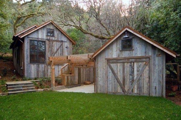 Rustic wooden shed with barn-style doors and pergola in a wooded backyard landscape.