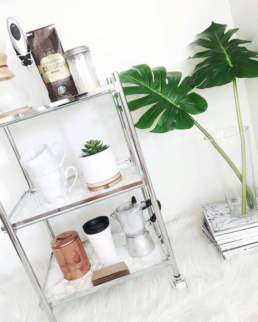 Modern metal bar cart with coffee accessories, mugs, and a potted succulent next to tropical leaves.