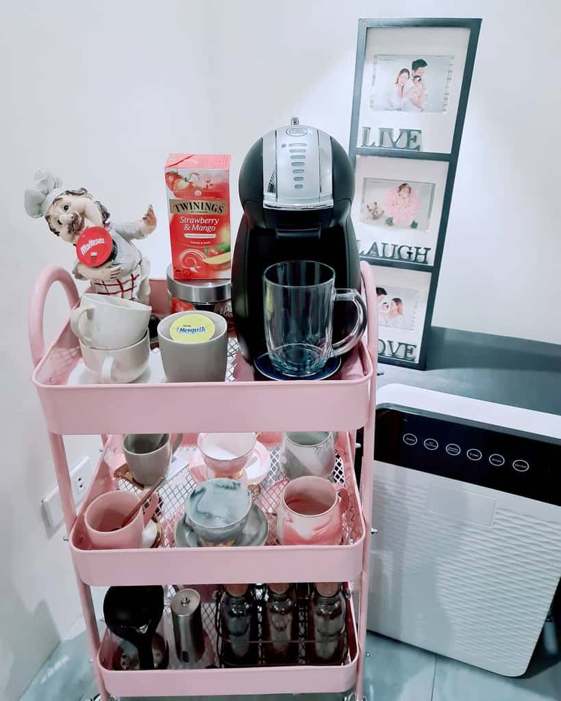 Pink tea and coffee cart with cups, tea boxes, coffee machine, and decorative figurine.