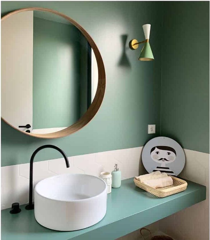 Modern bathroom with round mirror, green walls, white sink, black faucet, and decorative items on a shelf