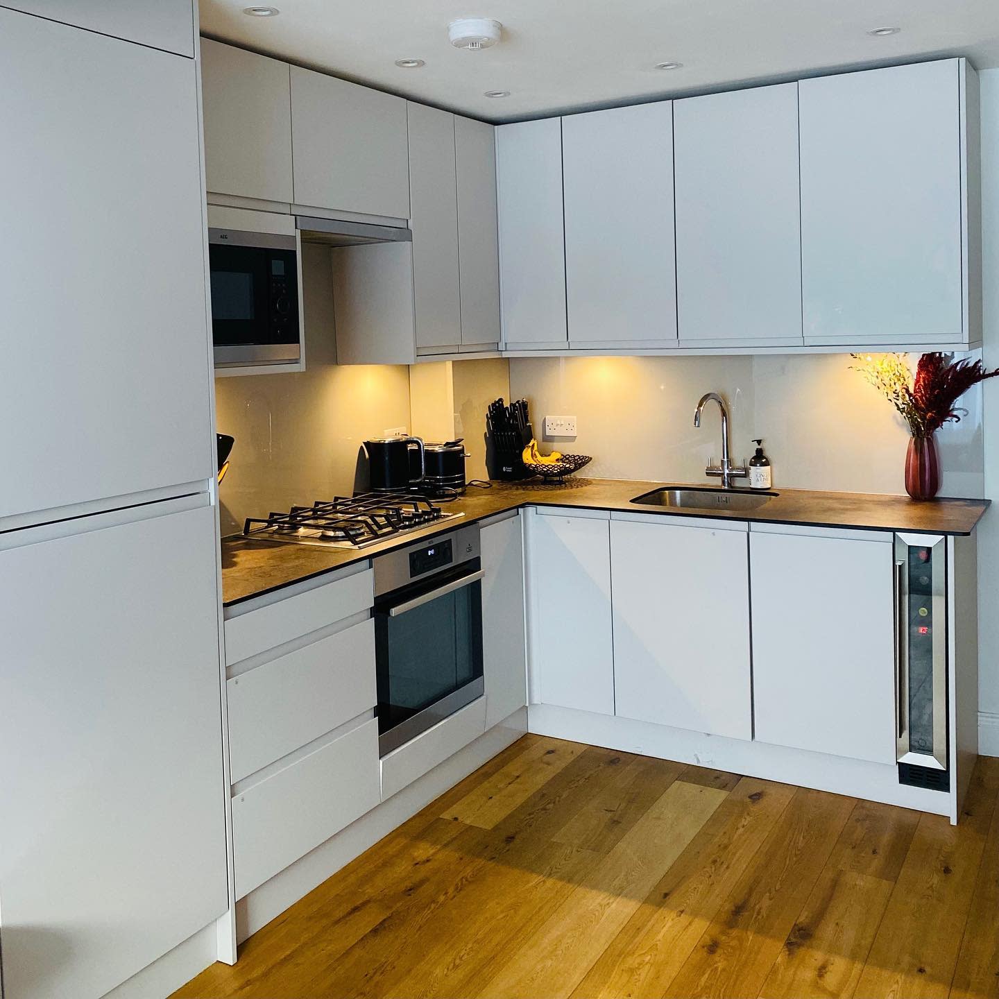 Modern white kitchen with built-in appliances, wooden flooring, and under-cabinet lighting.