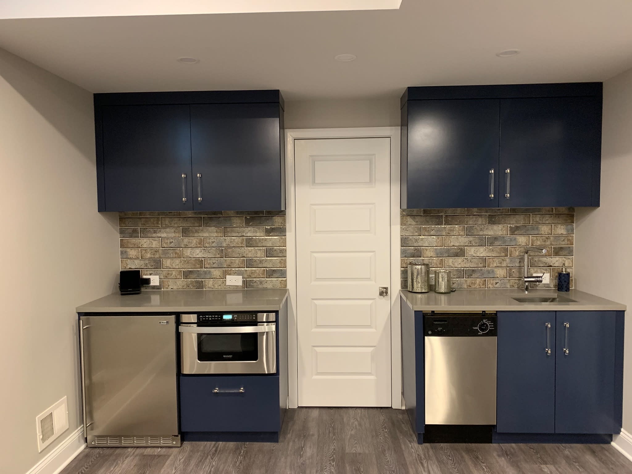 Small compact kitchen with blue cabinets, stainless steel appliances, and brick-style backsplash.