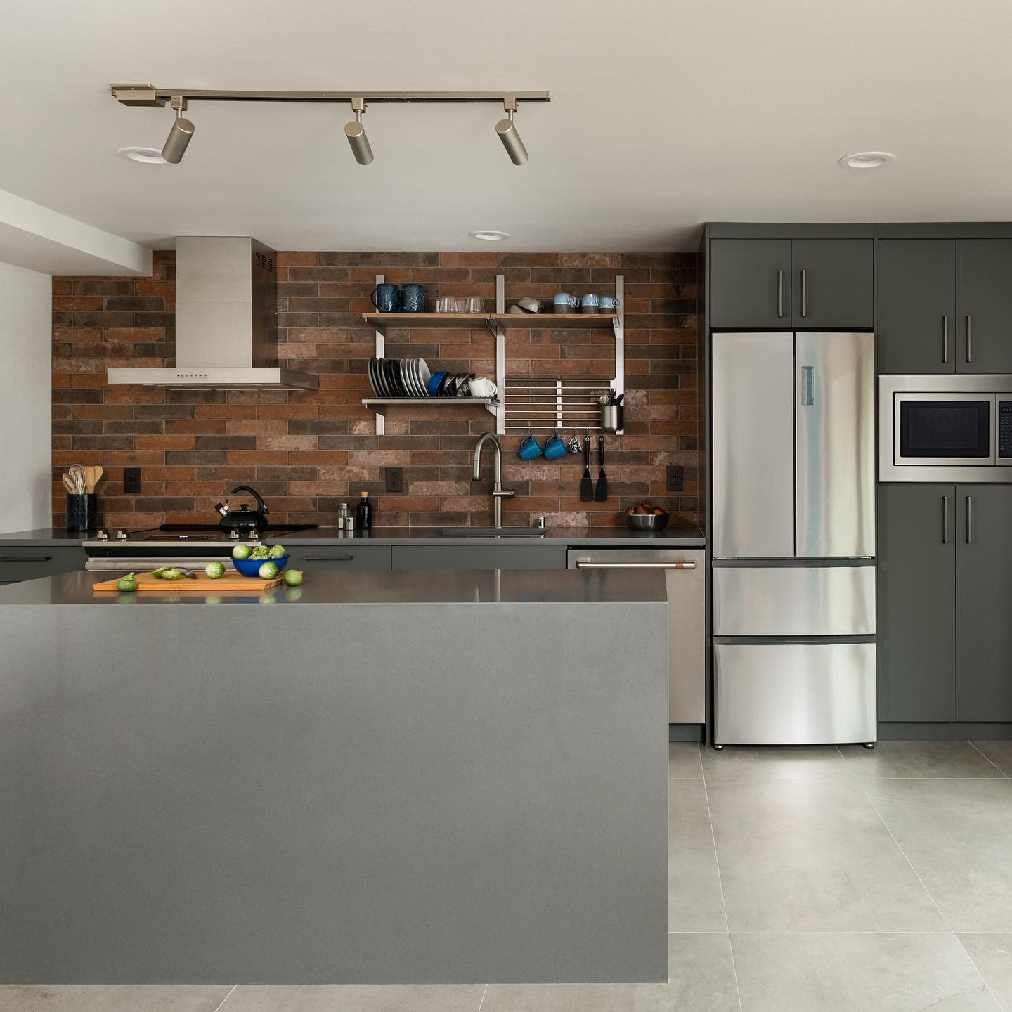 Modern kitchen with large grey island, brick backsplash, and open shelving for dishes.