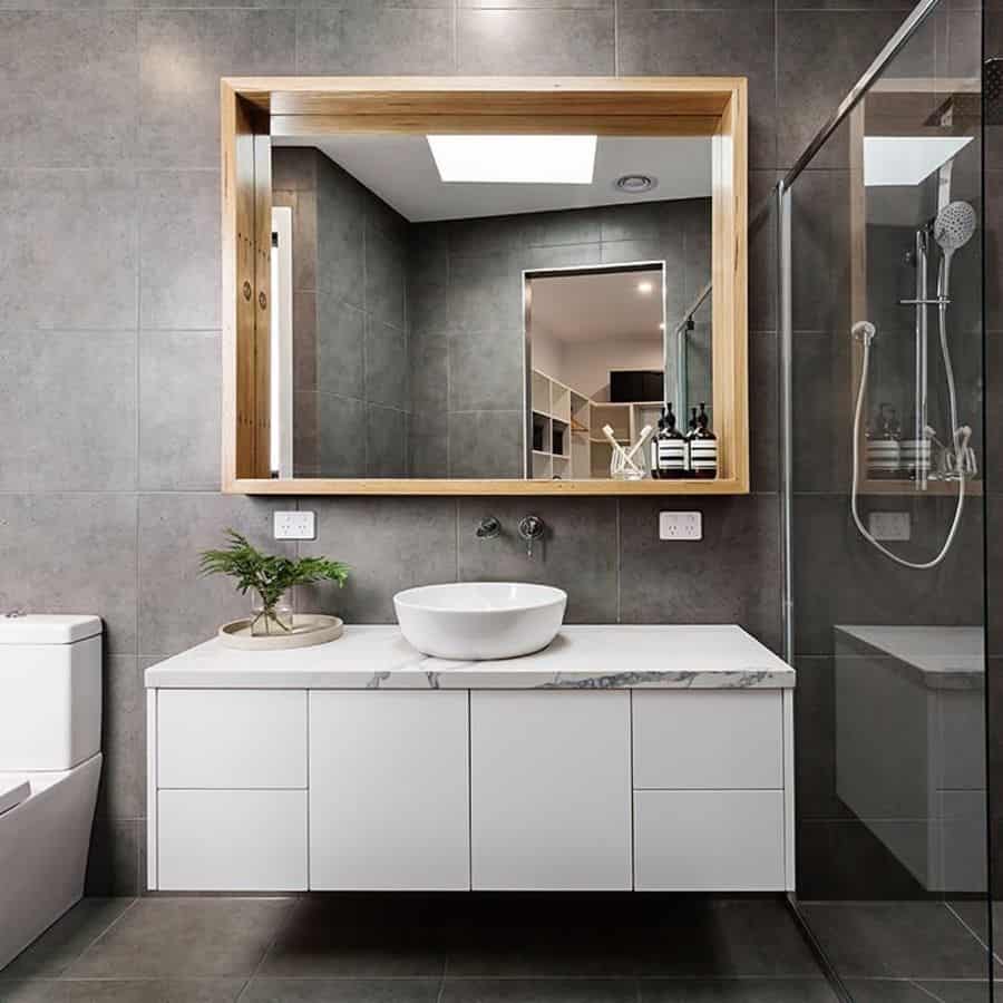 Modern bathroom with grey tile walls, wooden framed mirror, white floating vanity, and glass shower.