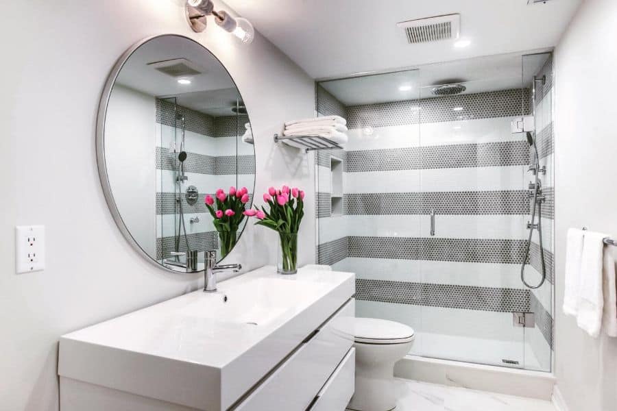 White contemporary bathroom with round mirror, striped shower tiles, and vase of pink tulips.