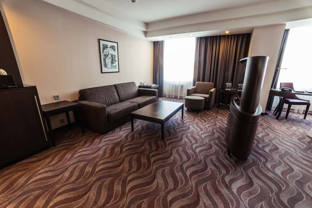 Brown hotel room with a sofa, coffee table, armchair, and wavy-patterned carpet—large window with curtains