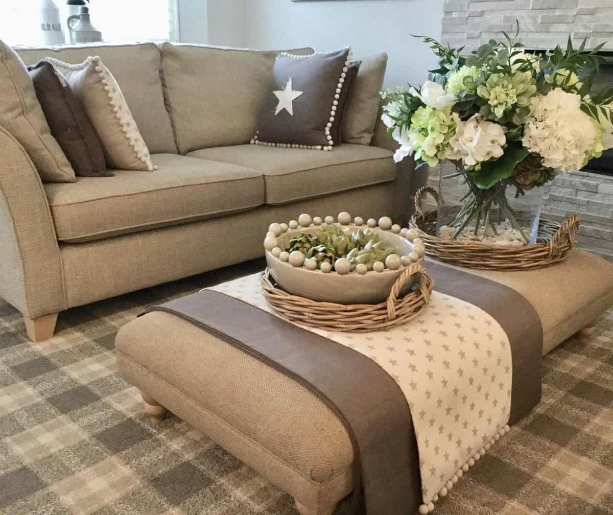 Cozy living room with a beige sofa, brown patterned ottoman, woven baskets, and a vase of white flowers