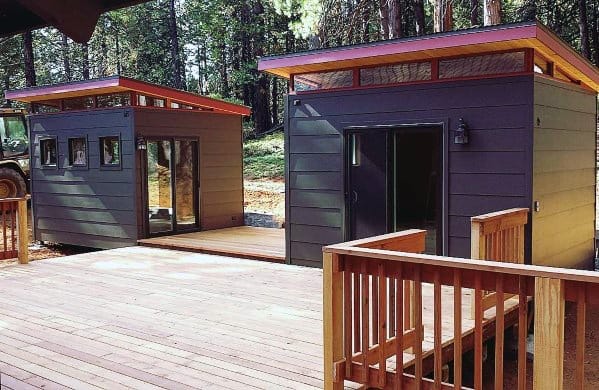 Two sleek grey backyard sheds with slanted roofs and wooden decks in a wooded area.