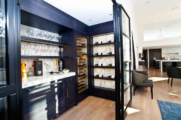 Luxury home bar with dark wood cabinets, marble accents, wine fridge, and glass shelving.