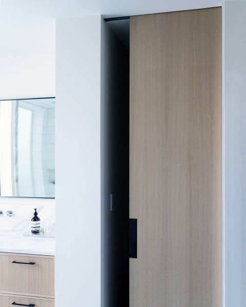 Modern wood pocket door with sleek black handle in a minimalist bathroom featuring a white countertop and neutral tones