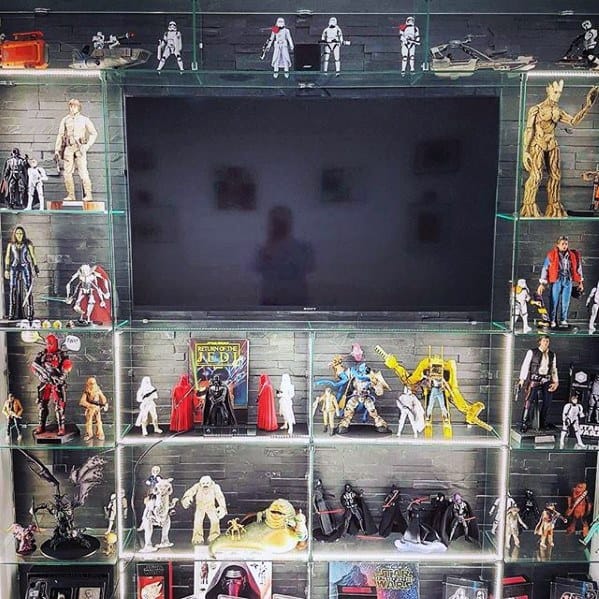 Display of various action figures and collectibles around a mounted TV on a brick wall with glass shelves