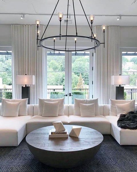 Modern living room with white sectional sofa, round wooden coffee table, large windows, and a circular chandelier