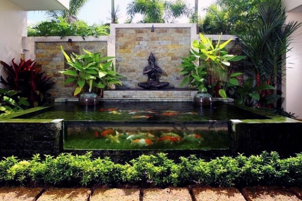 Raised koi pond with a glass window, surrounded by lush plants and featuring a meditative statue backdrop.