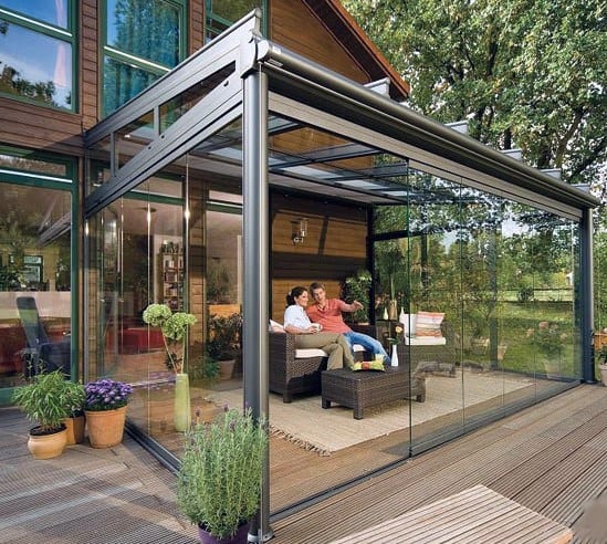 A modern glass-enclosed sunroom with sleek black frames, wicker seating, and potted plants.