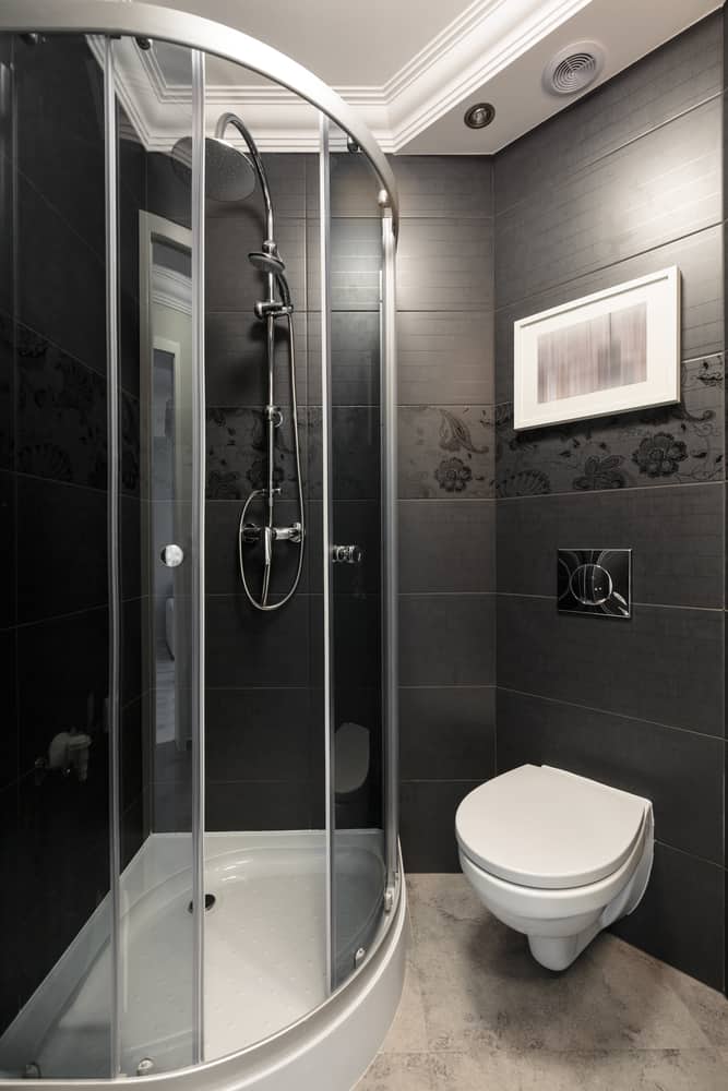 Small modern bathroom with black walls, corner glass shower, wall-mounted toilet, and decor.