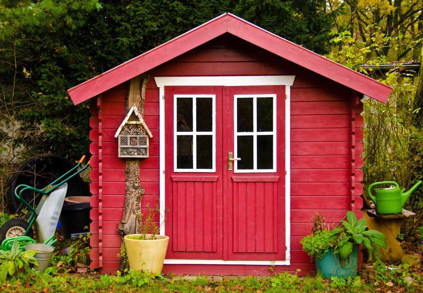 Small red wooden garden shed with double doors and potted plants in a lush backyard.