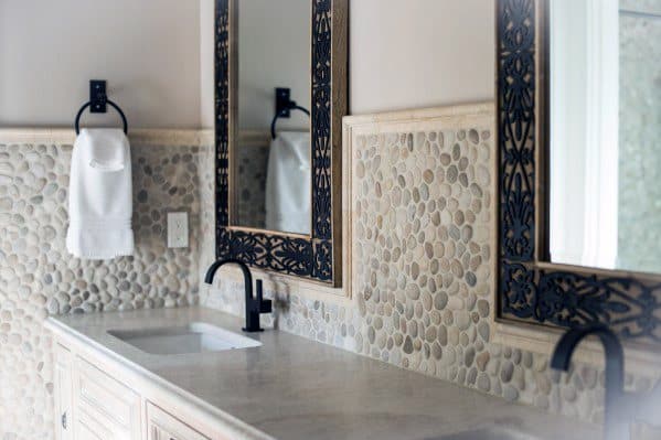 Bathroom with pebble tile backsplash, black faucets, and carved wooden mirrors.