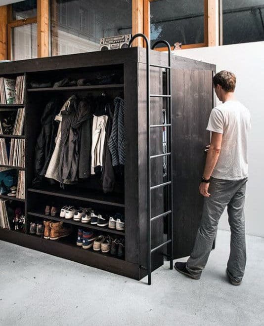 A man stands beside a large black shelving unit with clothes, shoes, and a ladder attached