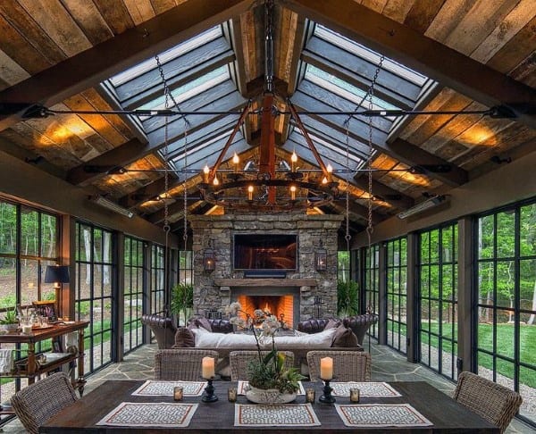 A rustic sunroom with a stone fireplace, wooden ceiling, glass walls, and cozy wicker seating.