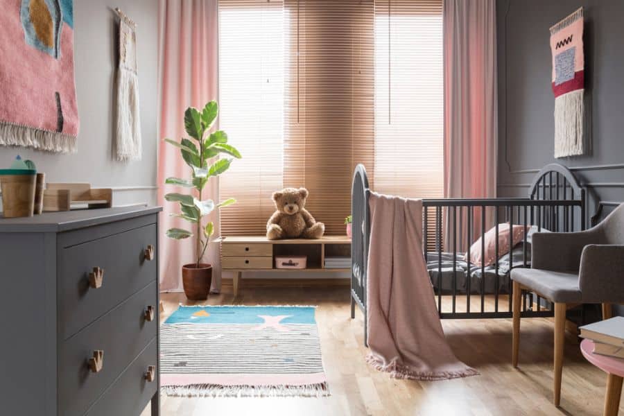 Moody baby room with dark crib, gray dresser, woven wall decor, and soft pink accents.