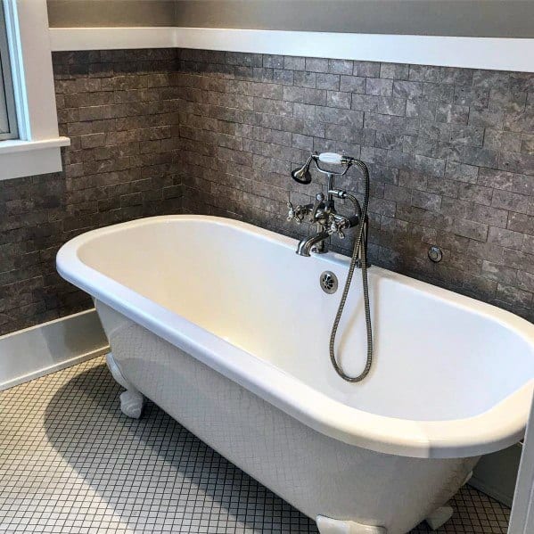 Freestanding white bathtub with vintage faucet and stone-tiled wall backdrop.