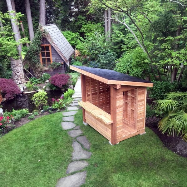 Wooden outdoor bar shed with a slanted roof in a lush garden with a stone path.