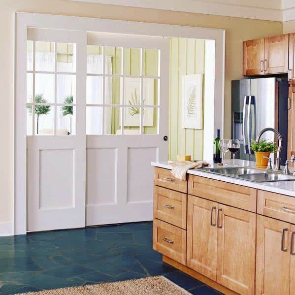 White pocket doors with glass panels divide a bright kitchen and sunroom, featuring natural wood cabinets and green accents