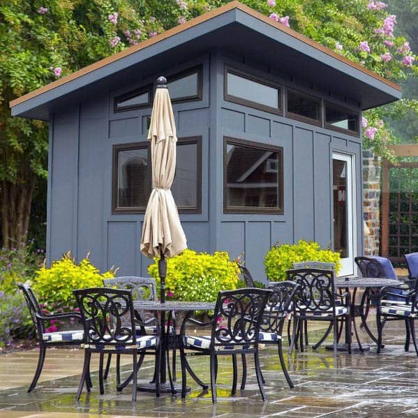 Contemporary blue shed with a slanted roof, large windows, and an outdoor patio set.