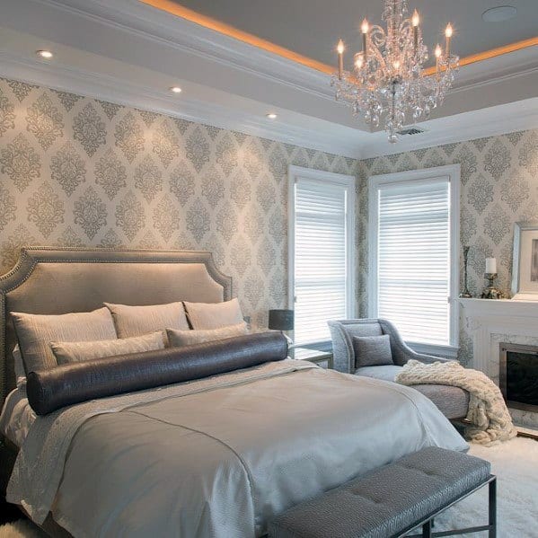 Elegant bedroom with gray damask wallpaper, tufted bed, chandelier, and cozy seating area.