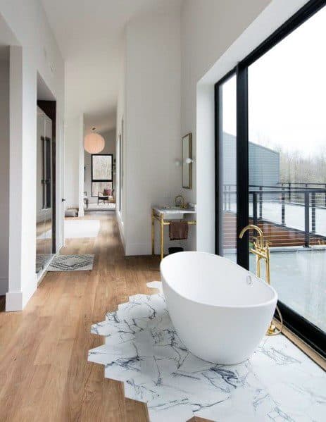 Freestanding bathtub by large windows with a wood and marble tile floor transition.