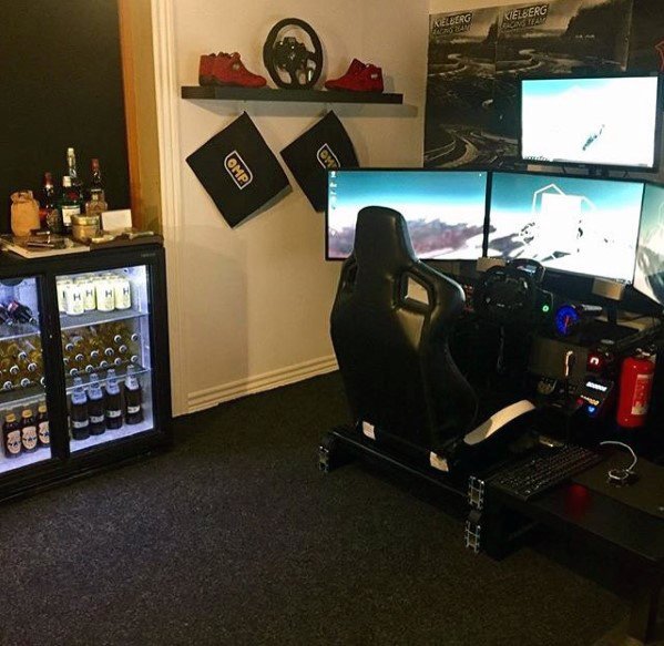 Sim racing setup with three monitors, racing seat, and steering wheel; mini fridge and shelves in the background