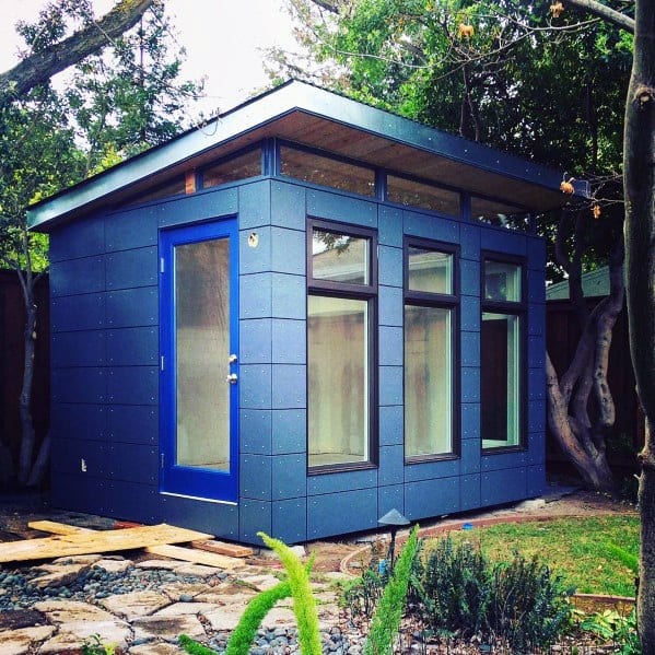 Modern aluminum shed with large windows and a blue door, set in a landscaped backyard.