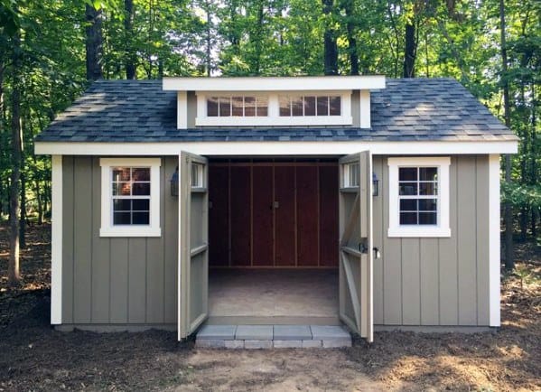 Pre-fabricated vinyl shed with double doors and dormer windows in a forested backyard.