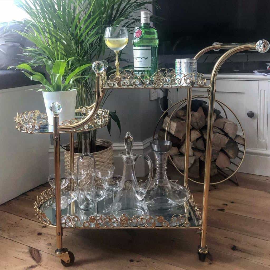 Elegant gold bar cart with crystal decanters, glassware, and a gin bottle next to potted plants.