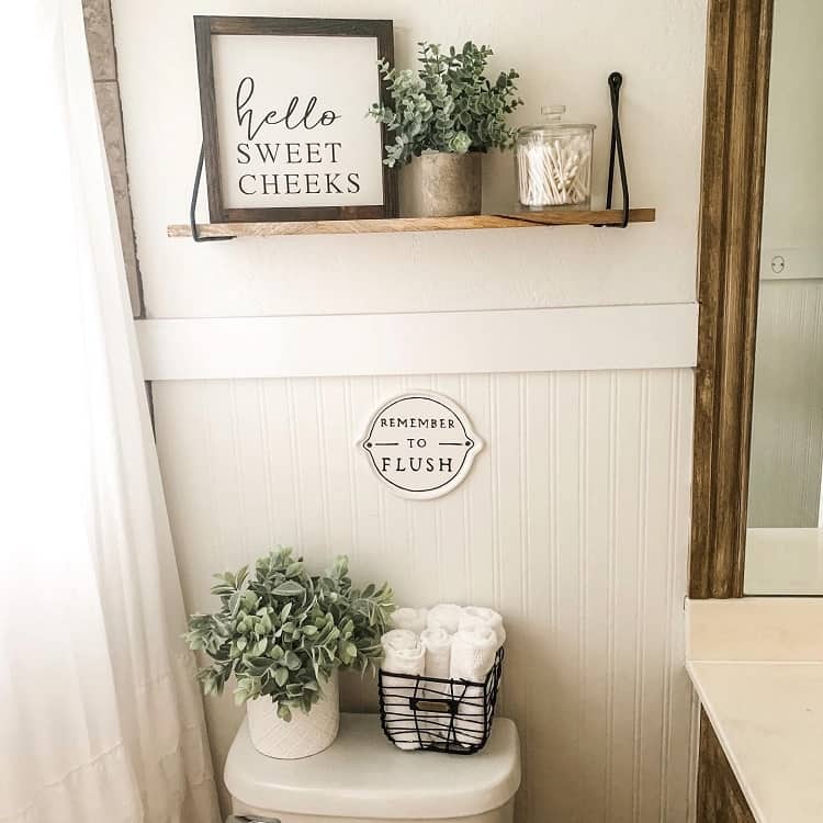 Farmhouse bathroom with floating shelf, potted plants, "Hello Sweet Cheeks" sign, and towel basket.