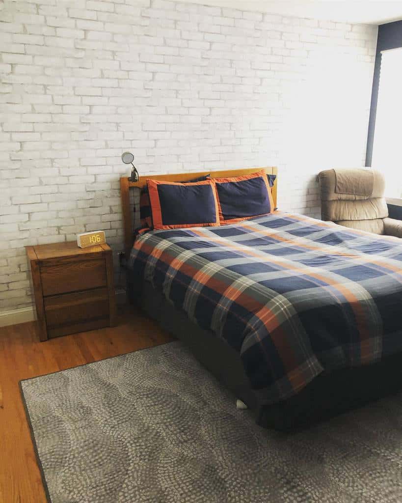 Bedroom with white brick wall, plaid bedding, wooden nightstand, and gray textured rug.