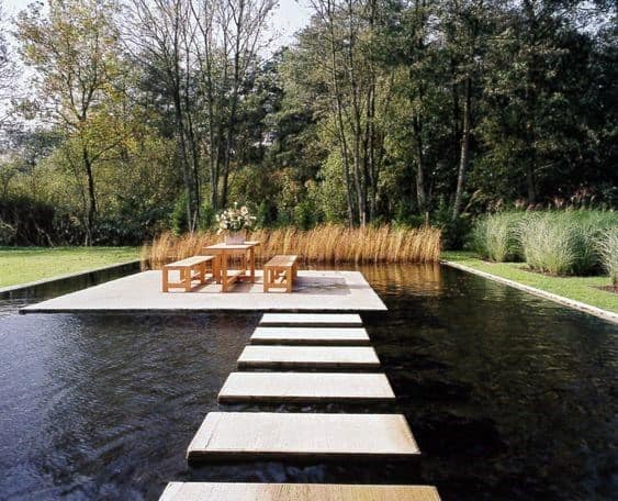 Floating stone steps leading to a wooden dining area on a platform, surrounded by a tranquil pond.