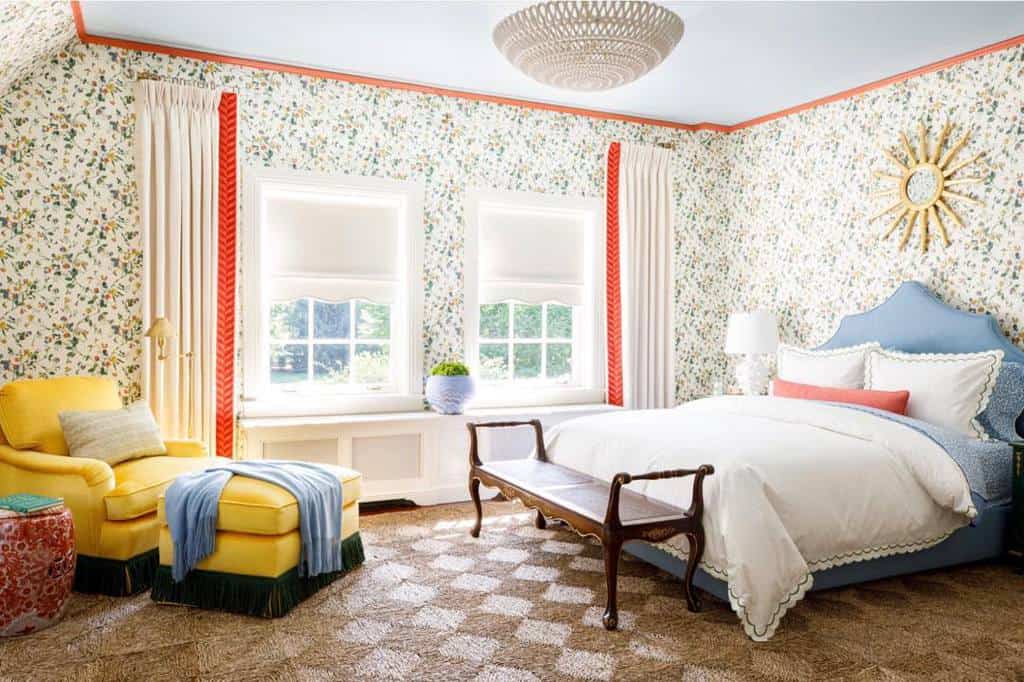 Bedroom with floral wallpaper, yellow armchair, blue upholstered bed, and woven rug.