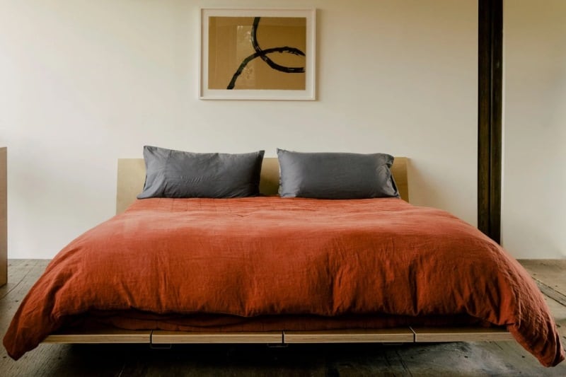 Minimalist bedroom with wooden platform bed, rust-colored duvet, and abstract wall art.