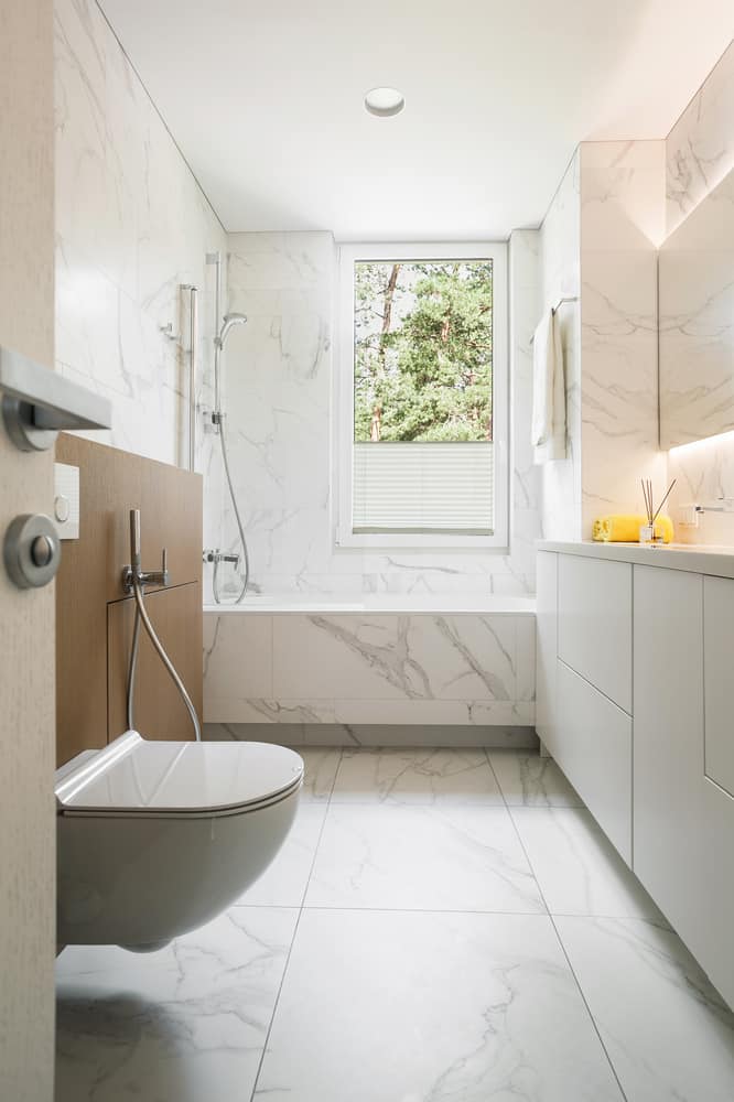 Bright bathroom with white marble tiles, floating toilet, bathtub, and large window.