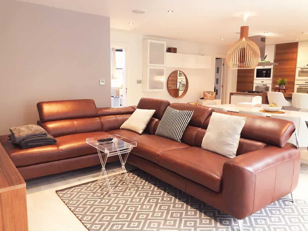 Living room with a stylish brown leather sectional, pillows, glass table, and patterned rug; modern kitchen in the background