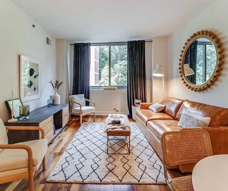 Cozy living room with brown leather sofa, patterned rug, round mirror, and large window with dark curtains