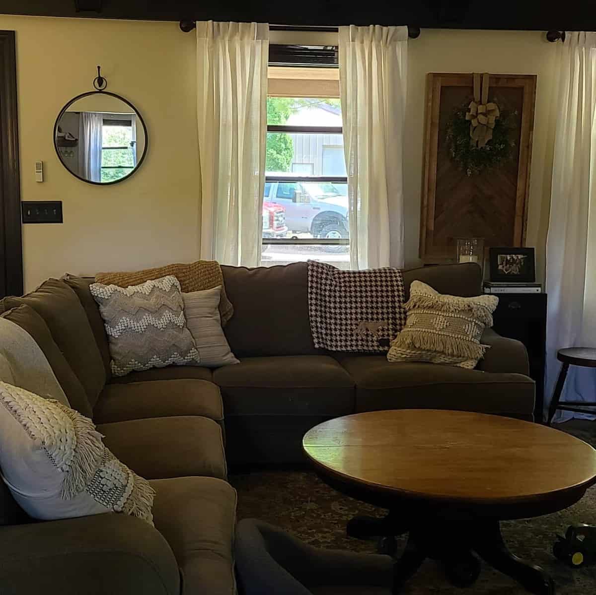 Cozy living room with a gray sectional, brown decorative pillows, a round mirror, and a window with white curtains