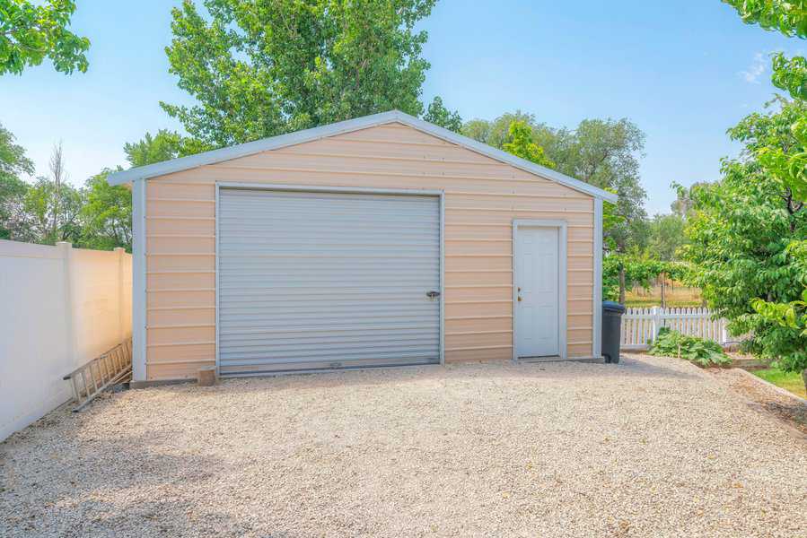 Beige garage-style shed with a large rolling door and side entry door, set in a gravel driveway.