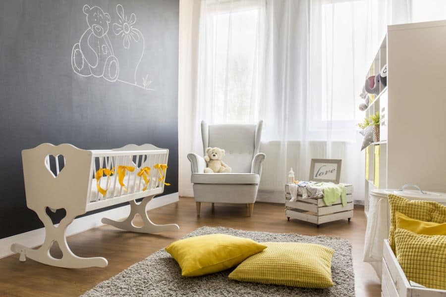 Charming nursery with a wooden rocking cradle, gray armchair, yellow cushions, and a chalkboard wall.