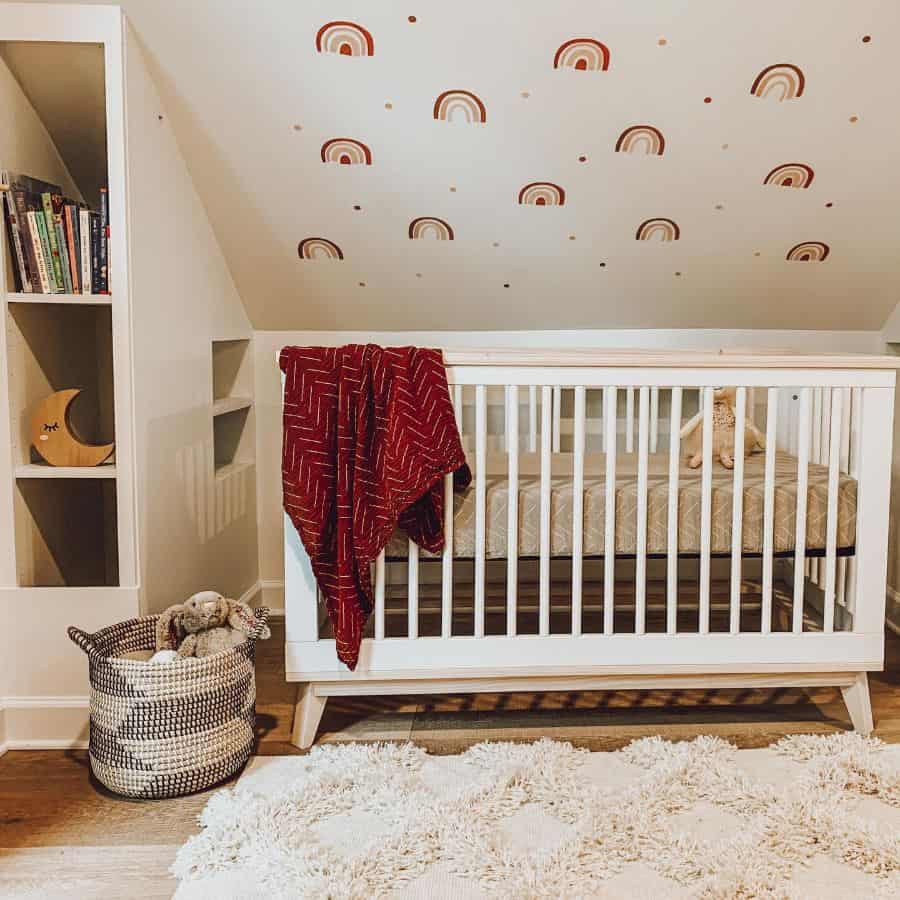 White crib with rainbow wall decals, cozy red blanket, and soft textured rug in an attic nursery.